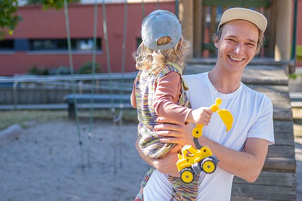 Das Bild zeigt einen Mitarbeiter, der auf einem Spielplatz sein Kind auf dem Arm hält